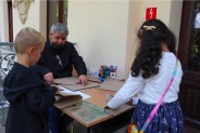 Mężczyzna siedzący przy stoliku oraz dwójka dzieci stojących przed nim i wykonujących samoloty z…