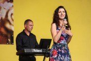 Na pierwszym planie Aleksandra Turek w kwiecistej sukni z mikrofonem w dłoni. Z tyłu jeden z…