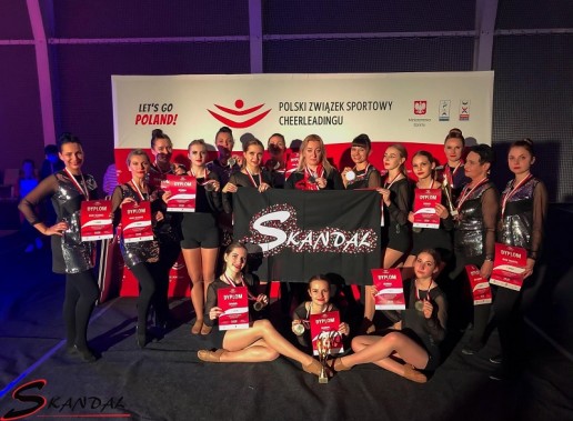 16 cheerleaderek z medalami, dyplomami, pucharem, trenerką trzymająca flagę z napisem Skandal
