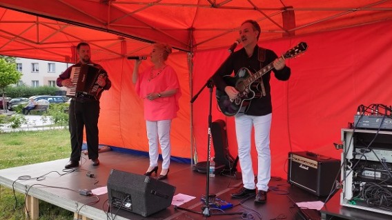 Dwoje mężczyzn - jeden z akordeonem, drugi z gitarą i jedna kobieta śpiewająca do mikrofonu na…
