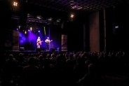 Widok ogólny, od tyłu na salę podczas koncertu. Na widowni siedzą ludzie. Na scenie w niebieskim…