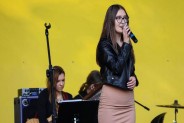Na pierwszym planie wokalistka Zuzanna Krupa. Ma długie, ciemne włosy i okulary. Ubrana jest w…
