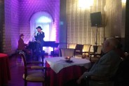 Na zdjęciu widoczny pianista w kapeluszu grający na fortepianie, oraz stojący wokalista ubrany w…